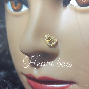 May include: A gold nose ring with a heart-shaped design and a bow in the center. The ring is adorned with small, clear rhinestones. The text "Heart bow" is written below the ring. The text "Pink Glam Shop" is written at the bottom of the image with a pink heart.