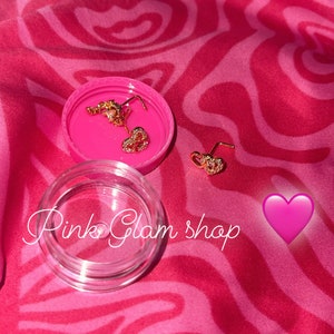 May include: Two gold heart-shaped nose rings with clear rhinestones, in a pink container with the text "Pink Glam Shop".