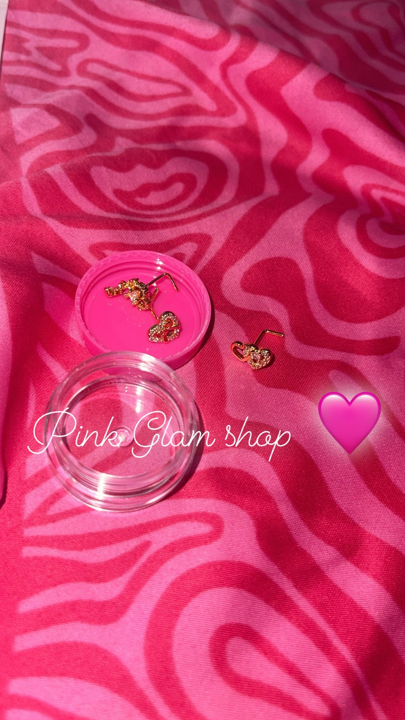 May include: Two gold heart-shaped nose rings with clear rhinestones, in a pink container with the text "Pink Glam Shop".