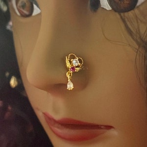 May include: A gold nose ring with a heart-shaped design and a dangling pearl. The ring is adorned with clear and pink gemstones.