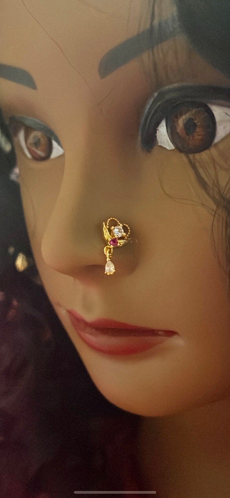 May include: A gold nose ring with a heart-shaped design and a dangling pearl. The ring is adorned with clear and pink gemstones.