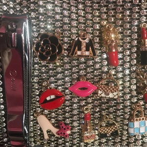 May include: A close-up of a variety of charms, including a black and white Chanel-inspired charm, a red lipstick charm, a red and white striped charm, and a black and white checkered charm. The charms are all attached to a silver chain.