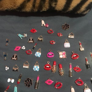 May include: Assortment of colorful enamel charms featuring various fashion and beauty themed designs.  The charms include lips, purses, shoes, lipstick, wine glasses, and makeup palettes.  The charms are all gold-toned and have a shiny finish.