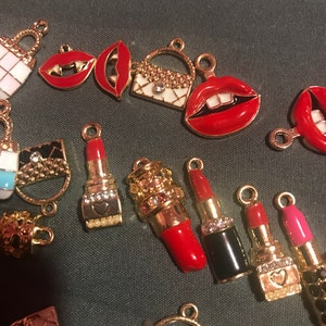 May include: A collection of gold-tone charms featuring red lips, lipstick tubes, and small purses. The charms are decorated with rhinestones and enamel.