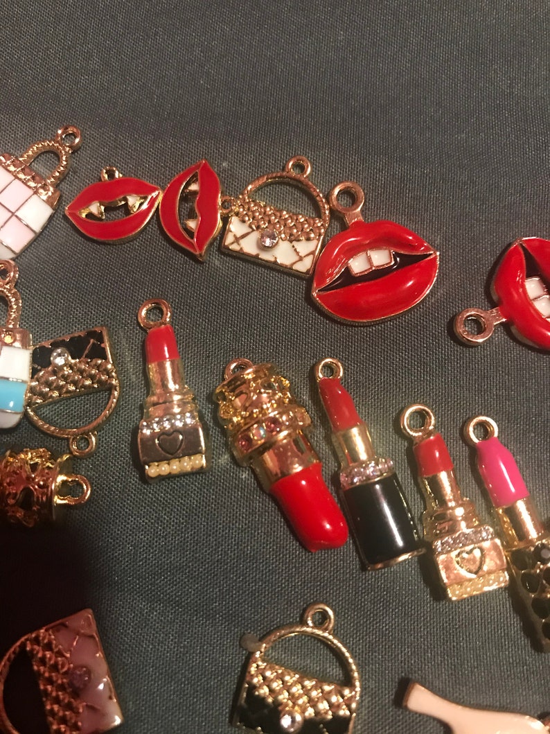 May include: A collection of gold-tone charms featuring red lips, lipstick tubes, and small purses. The charms are decorated with rhinestones and enamel.