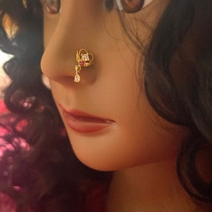 May include: A gold nose ring with a red stone and clear rhinestones. The nose ring is worn by a person with dark hair.