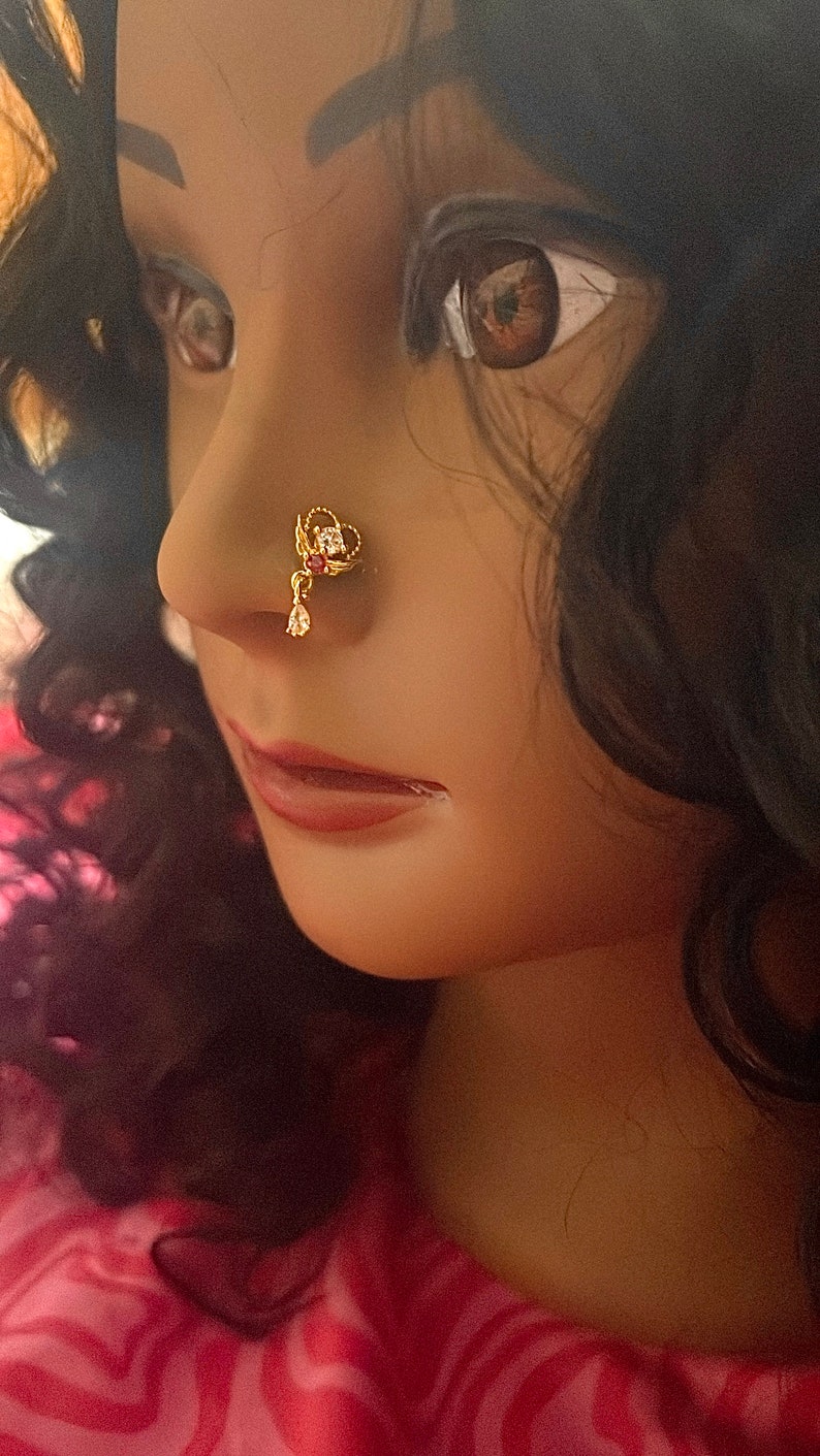 May include: A gold nose ring with a red stone and clear rhinestones. The nose ring is worn by a person with dark hair.