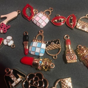 May include: A collection of gold-tone charms featuring various fashion-themed designs, including lips, handbags, shoes, and a flower. The charms are decorated with enamel in various colors, including red, pink, white, blue, and black.
