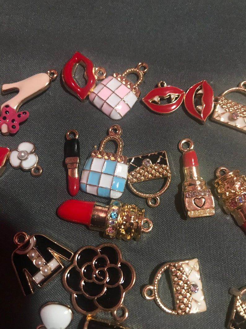 May include: A collection of gold-tone charms featuring various fashion-themed designs, including lips, handbags, shoes, and a flower. The charms are decorated with enamel in various colors, including red, pink, white, blue, and black.