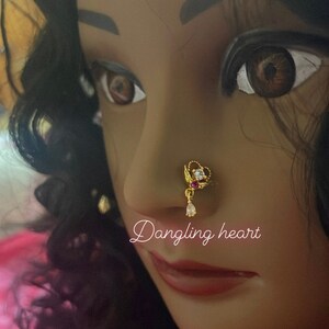 May include: A gold nose ring with a dangling heart charm and clear and pink stones. The text "Dangling heart" is written below the nose ring. The text "Pink Glam Shop" is written at the bottom of the image with a pink heart.