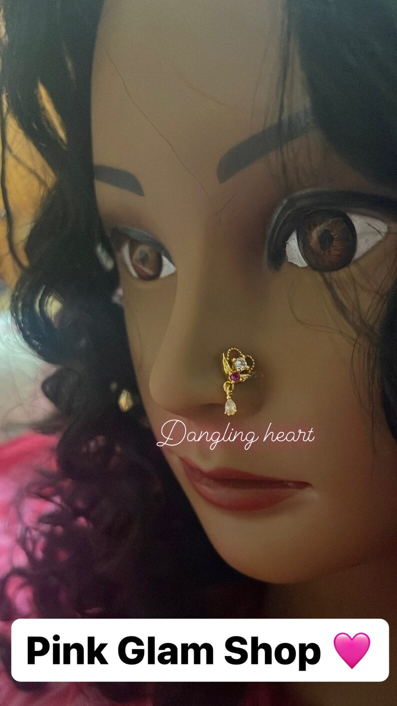 May include: A gold nose ring with a dangling heart charm and clear and pink stones. The text "Dangling heart" is written below the nose ring. The text "Pink Glam Shop" is written at the bottom of the image with a pink heart.