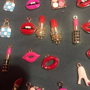 May include: Assortment of gold-tone charms with various makeup-themed designs, including lips, lipstick tubes, and a makeup palette. The charms are colorful and feature red, pink, and white enamel.
