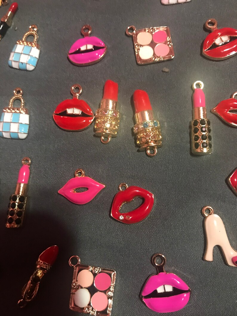 May include: Assortment of gold-tone charms with various makeup-themed designs, including lips, lipstick tubes, and a makeup palette. The charms are colorful and feature red, pink, and white enamel.