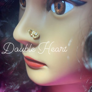 May include: A gold nose ring with two interlocking hearts and clear rhinestones. The nose ring is worn by a model with dark hair and red lipstick. The text "Double Heart" and "Pink Glam Shop" is visible in the image.