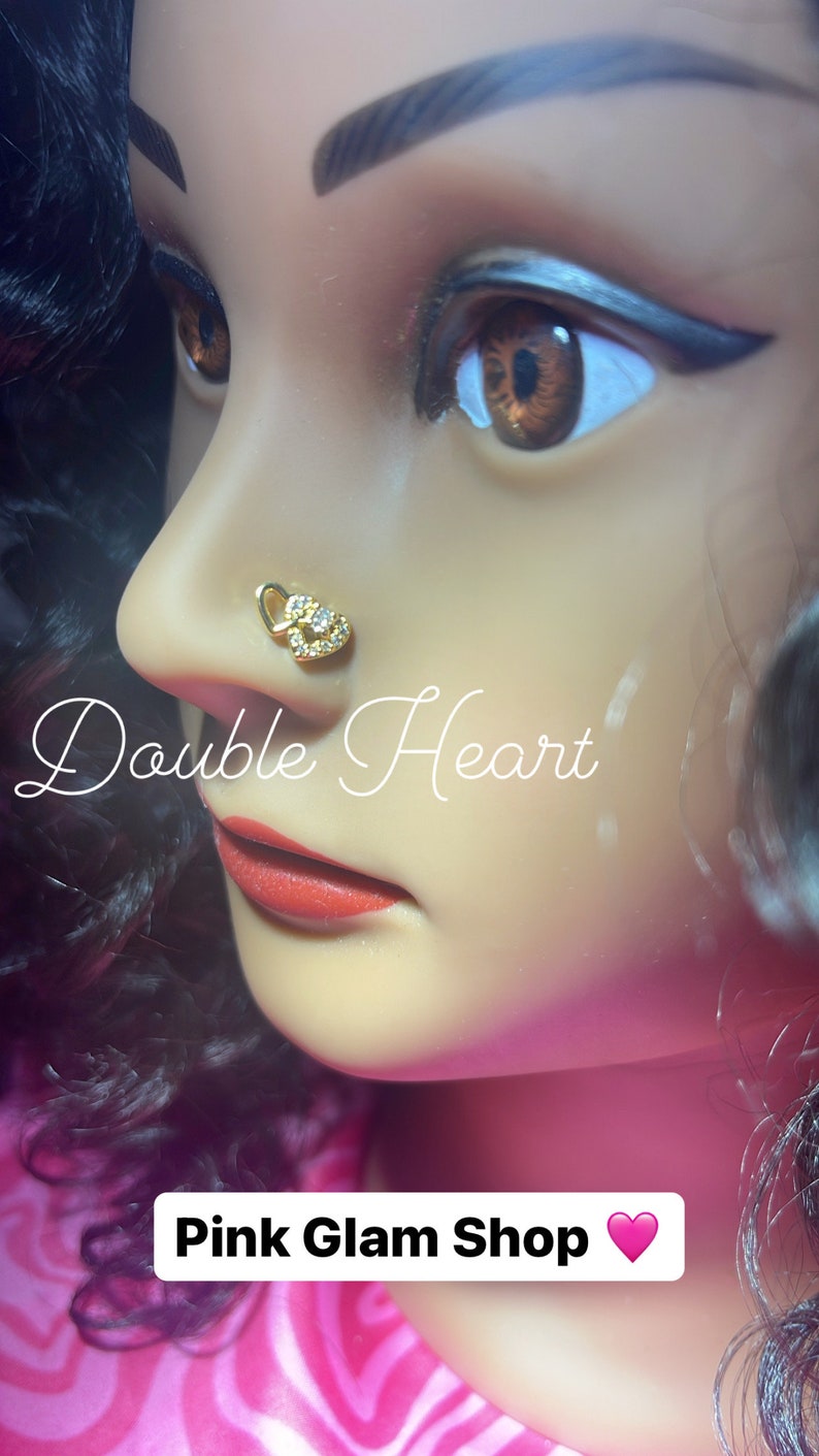 May include: A gold nose ring with two interlocking hearts and clear rhinestones. The nose ring is worn by a model with dark hair and red lipstick. The text "Double Heart" and "Pink Glam Shop" is visible in the image.