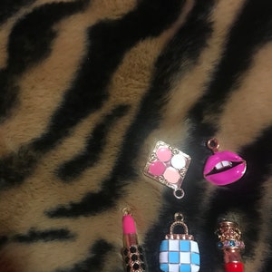 May include: Five gold-tone charms with a makeup theme. The charms include a lipstick, a makeup palette, a purse, a pair of lips, and another lipstick. The charms are on a brown and black fuzzy background.