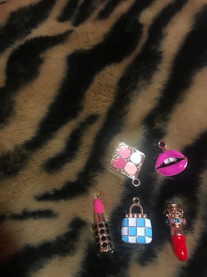 May include: Five gold-tone charms with a makeup theme. The charms include a lipstick, a makeup palette, a purse, a pair of lips, and another lipstick. The charms are on a brown and black fuzzy background.