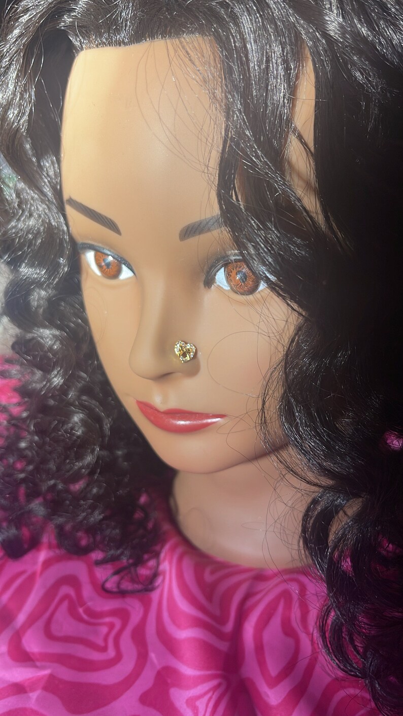 May include: A close-up of a mannequin's face with long, dark curly hair. The mannequin is wearing a gold heart-shaped nose ring.