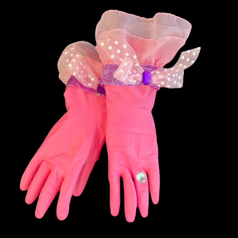 Pair of bright pink rubber cleaning gloves transformed into glamorous fashion accessories. Long gloves feature voluminous sheer pink polka-dot ruffle cuffs with glittery purple ribbon bows, purple gem accents, and a sparkling crystal ring on one finger. Playful, bold, and feminine style.