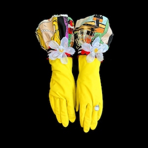 May include: A pair of bright yellow rubber gloves featuring ruffled cuffs with a colorful comic book style print. Each glove is adorned with a white fabric flower at the wrist, and one glove features a large, clear, faceted faux gemstone ring.