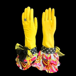 May include: A pair of bright yellow rubber gloves featuring decorative cuffs. The cuffs are adorned with a black and white polka dot band, a black fabric flower, and a ruffled hem in a vibrant pink, yellow, and white paisley pattern.