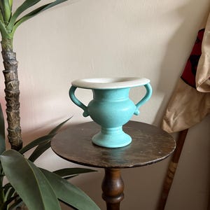 May include: A turquoise ceramic vase with a white rim and two handles. The vase is displayed on a small, dark wooden table. The vase has a pedestal base and a classic urn shape. The background includes a plant and a wall.