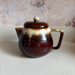 May include: A round ceramic teapot featuring a glossy dark brown base that transitions into a creamy white drip glaze near the top. The teapot includes a matching lid with a small knob handle and a curved side handle, showcasing a smooth finish.