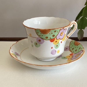 May include: A white porcelain teacup and saucer set featuring a hand-painted pattern of colourful abstract circles, dots, and floral shapes in shades of orange, green, purple, and yellow, with a scalloped rim and a delicate handle on the cup.