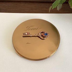 May include: A round gold-toned compact mirror featuring a decorative key emblem on the lid. The key is embellished with small blue, orange, and clear stones. The word "Jemi" is engraved in cursive script above the key on the metallic surface.