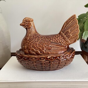Peut inclure: Un plat en céramique émaillée marron en forme de poule sur un nid. La poule est représentée en position assise avec des textures de plumes détaillées, reposant sur une base moulée pour ressembler à un panier tressé avec une finition brillante.