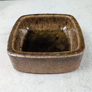 Peut inclure: Un petit plat carré en céramique avec un glaçage brun texturé. Le plat a des coins arrondis et un bord légèrement surélevé. L'intérieur est d'une nuance de brun plus foncée et le plat semble vide. Le plat est probablement utilisé pour servir de petites portions de nourriture ou comme objet décoratif.
