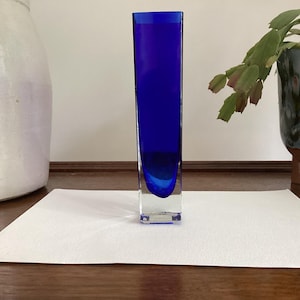 May include: A tall, rectangular glass vase featuring a deep cobalt blue colour. The base of the vase is clear, transitioning into a solid blue body with a smooth, polished finish. The design is vertical and slender, with a flat top opening.