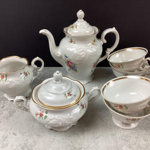 May include: A white porcelain tea set with floral accents. The set includes a teapot, sugar bowl, creamer, and two teacups with saucers. Gold trim edges the cups and sugar bowl. The set is on a gray surface.