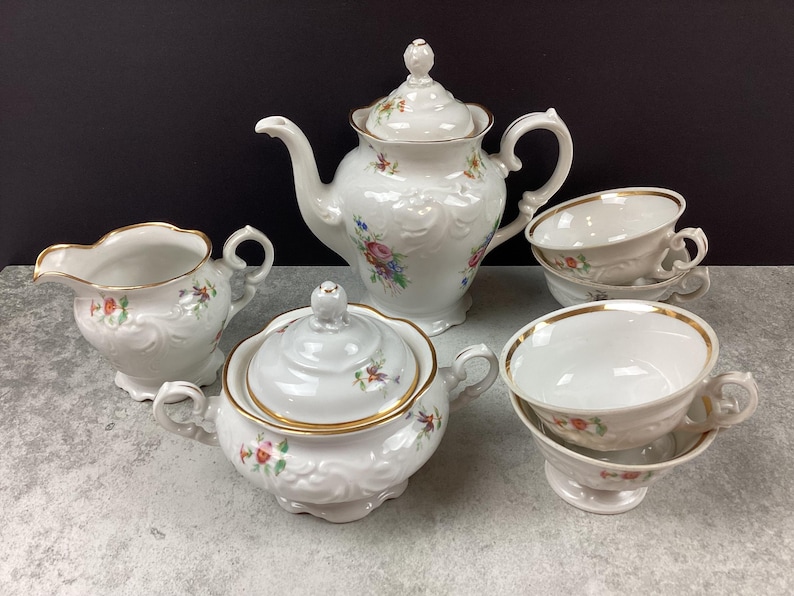 May include: A white porcelain tea set with floral accents. The set includes a teapot, sugar bowl, creamer, and two teacups with saucers. Gold trim edges the cups and sugar bowl. The set is on a gray surface.