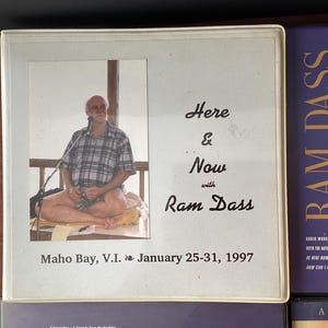 May include: A white framed photo of Ram Dass sitting cross-legged on a cushion. The photo is titled "Here & Now with Ram Dass" and includes the text "Maho Bay, V.I. January 25-31, 1997".