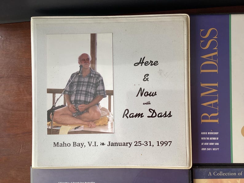 May include: A white framed photo of Ram Dass sitting cross-legged on a cushion. The photo is titled "Here & Now with Ram Dass" and includes the text "Maho Bay, V.I. January 25-31, 1997".