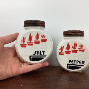 以下が含まれることがあります： ヴィンテージの白い塩とコショウ入れのペア。各シェーカーは、"SALT"と"PEPPER"の文字が入った赤と黒のデザインです。蓋は錆びた茶色です。シェーカーは側面にテクスチャ加工されたリブのデザインが施されています。