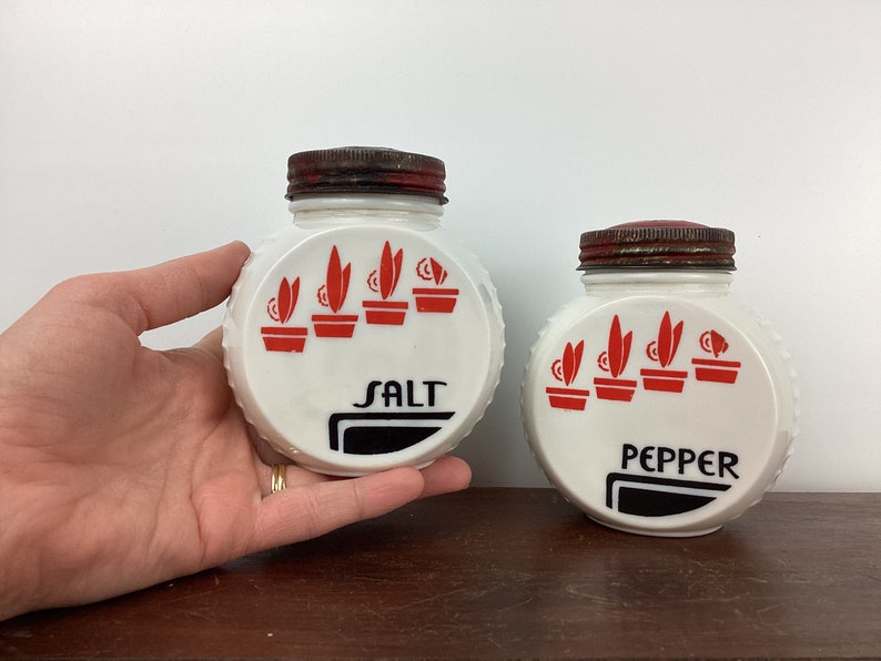 以下が含まれることがあります： ヴィンテージの白い塩とコショウ入れのペア。各シェーカーは、"SALT"と"PEPPER"の文字が入った赤と黒のデザインです。蓋は錆びた茶色です。シェーカーは側面にテクスチャ加工されたリブのデザインが施されています。