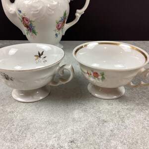 May include: Two white porcelain teacups with gold trim and floral accents. The teacups have a pedestal base and curved handles. A white teapot with floral designs is visible in the background, suggesting a tea set.