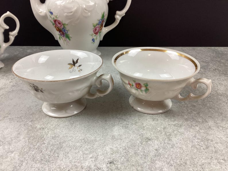 May include: Two white porcelain teacups with gold trim and floral accents. The teacups have a pedestal base and curved handles. A white teapot with floral designs is visible in the background, suggesting a tea set.