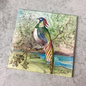 May include: Square, hand-painted artwork featuring a colorful bird perched on a branch. The bird has a blue head, green and white body, and a long, striped tail. The background depicts a landscape with a river and foliage. Signed "Rola" in the bottom right corner.
