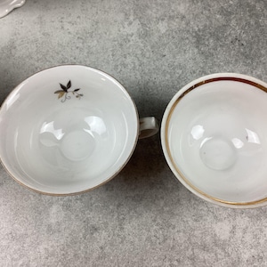 May include: Two white porcelain teacups with gold rims. One cup has a small floral design in gold and black. The cups are on a gray surface. The teacups have small handles.