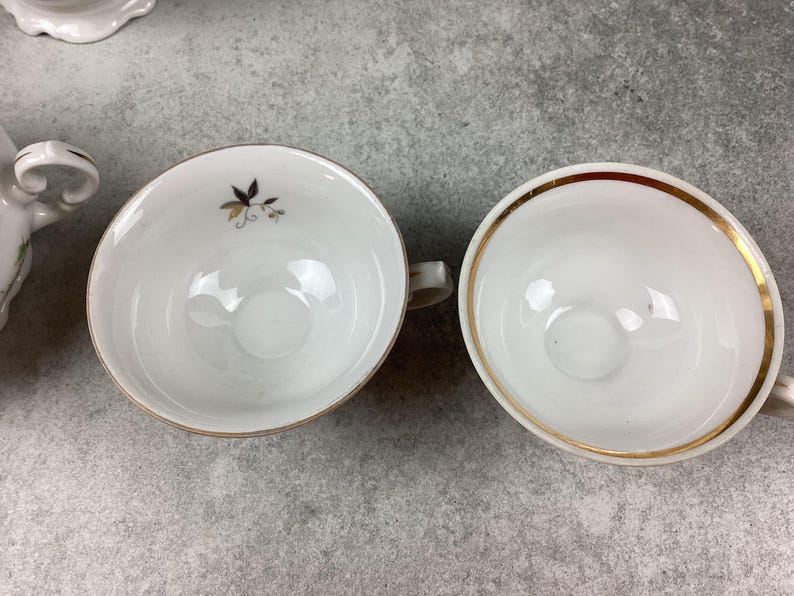 May include: Two white porcelain teacups with gold rims. One cup has a small floral design in gold and black. The cups are on a gray surface. The teacups have small handles.