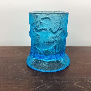 Könnte beinhalten: Ein blauer Glasbecher mit strukturierter Oberfläche und einem Design einer Figur in einem Baum. Der Becher ist perfekt für die Aufbewahrung von Zahnbürsten, Stiften oder anderen kleinen Gegenständen.