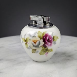 以下が含まれることがあります： ピンク、紫、黄色のバラをあしらった花柄の、ヴィンテージの白いセラミックライター。ライターは銀色の金属製のトップが付いています。ライターは大理石の表面にあります。
