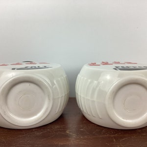 以下が含まれることがあります： 樽のデザインの白いセラミック製の塩とコショウ入れが2つ。片方のシェーカーには「SALT」の文字が黒と赤で印刷され、もう片方には「PEPPER」の文字が印刷されています。シェーカーは茶色の表面にあります。