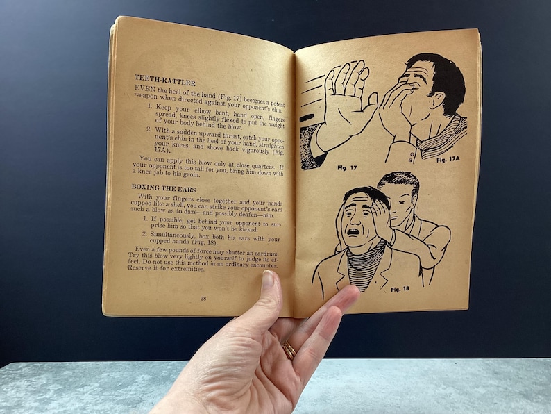 May include: Open book with illustrations and text on self-defense techniques. The illustrations depict hand gestures and facial expressions. The text includes instructions for various moves, such as the "Teeth-Rattler" and "Boxing the Ears."
