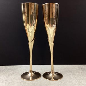 Puede incluir: Dos copas de champán doradas con un diseño único. Las copas tienen un tallo delgado y una base ancha y redonda. El cristal se ensancha hacia la parte superior. El fondo es negro.