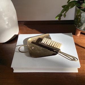 May include: A small, square brass crumb tray with a rounded handle and a matching brass-handled brush featuring light-colored bristles. The set is resting on a thick, white rectangular object, showcasing a metallic finish and functional design.