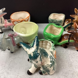 May include: A selection of ceramic donkey planters in various colours. The planters are designed as donkeys pulling carts, with the carts serving as containers for plants. The donkeys are painted in shades of brown, grey, and white.